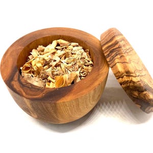 Könnte beinhalten: Ein hölzernes Mörser- und Stößel-Set. Der Mörser ist eine runde Schüssel, gefüllt mit getrockneten, hellbraunen Kräutern. Der Stößel hat eine zylindrische Form mit einer strukturierten Holzmaserung. Das Set ist aus Olivenholz.