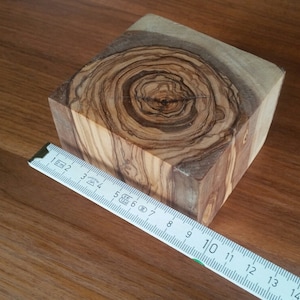 Könnte beinhalten: Eine Nahaufnahme eines quadratischen Holzblocks mit einem einzigartigen Maserungsmuster. Das Holz ist hellbraun mit dunkleren braunen Streifen und Wirbeln. Ein Lineal liegt neben dem Holzblock und zeigt, dass er etwa 20 cm breit ist.