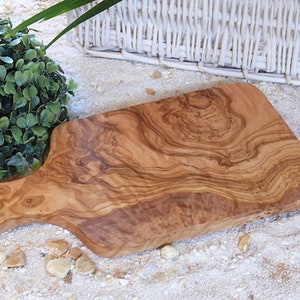 Pode incluir: Tábua de corte de madeira com uma pega, apresentando um padrão de grão de madeira natural. A tábua é castanho claro com redemoinhos castanhos escuros e um acabamento liso. A pega tem um orifício para pendurar. A tábua tem cerca de 30 cm de comprimento.