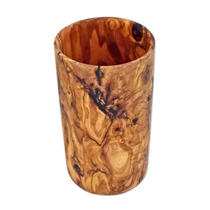 Puede incluir: Un vaso cil&iacute;ndrico de madera de olivo con un patr&oacute;n de vetas naturales. El vaso tiene un acabado liso y pulido. El interior es de un tono marr&oacute;n m&aacute;s oscuro. El vaso est&aacute; vac&iacute;o y sobre un fondo blanco.