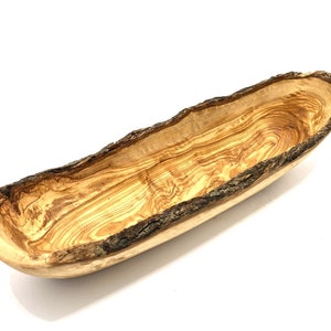 Può includere: Una lunga ciotola in legno d'ulivo con bordo naturale. La ciotola è marrone chiaro con venature più scure e un bordo di corteccia marrone scuro ruvido. L'interno è liscio e curvo.