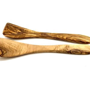 Könnte beinhalten: Zwei Holzspatel aus Olivenholz mit natürlicher Maserung. Die Spatel haben einen flachen, rechteckigen Kopf und einen langen, gebogenen Griff. Die Holzfarbe variiert von hell- bis dunkelbraun.