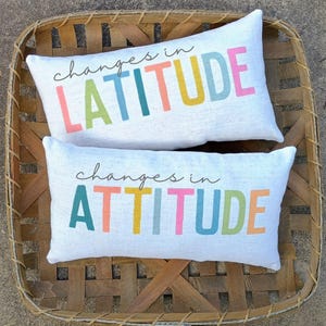 Pode incluir: Duas almofadas retangulares brancas com as frases "changes in LATITUDE" e "changes in ATTITUDE" impressas em letras coloridas. As almofadas estão dentro de uma cesta de vime.