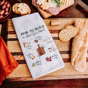 May include: A white kitchen towel with the text "How to build a charcuterie board" and a diagram showing different food items to include on a charcuterie board. The towel is on a wooden cutting board with a baguette, crackers, and olives.