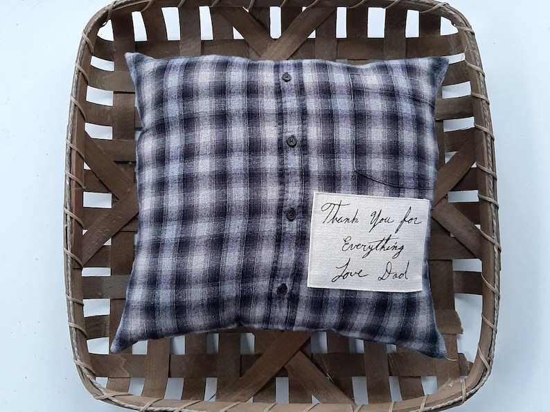 May include: A black and white plaid pillow with a white fabric patch sewn on the front. The patch has handwritten text that reads "Thank you for everything Love Dad". The pillow is sitting in a woven basket.