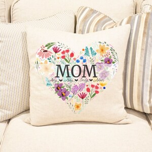Puede incluir: Una almohada blanca con un dise&ntilde;o de coraz&oacute;n floral. El coraz&oacute;n est&aacute; lleno de flores coloridas y la palabra "MOM" est&aacute; escrita en letras negras. Los nombres "Amy, Abby, Emily, Adam" est&aacute;n escritos en letras m&aacute;s peque&ntilde;as debajo de la palabra "MOM".