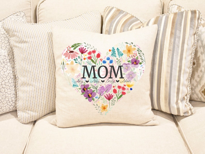 Puede incluir: Una almohada blanca con un dise&ntilde;o de coraz&oacute;n floral. El coraz&oacute;n est&aacute; lleno de flores coloridas y la palabra "MOM" est&aacute; escrita en letras negras. Los nombres "Amy, Abby, Emily, Adam" est&aacute;n escritos en letras m&aacute;s peque&ntilde;as debajo de la palabra "MOM".