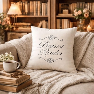 May include: A square, off-white pillow with the words "Dearest Reader" in elegant black script. The pillow is adorned with decorative flourishes above and below the text. It rests on a beige throw on a sofa, with a backdrop of books and a lamp.