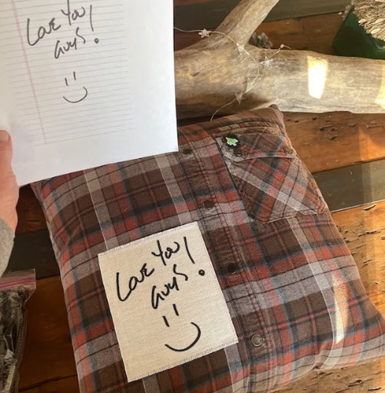 May include: A brown and red plaid pillow with a white patch sewn on the front. The patch has handwritten text that says "Love you! Guys." with a smiley face drawn below the text.