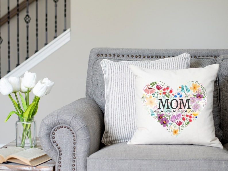 Puede incluir: Almohada blanca con un dise&ntilde;o de coraz&oacute;n floral y la palabra "MOM" en texto negro. El coraz&oacute;n est&aacute; rodeado de flores coloridas y los nombres "Amy, Ally, Emily, Alex" est&aacute;n escritos debajo de la palabra "MOM".