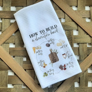 May include: White kitchen towel with a graphic design that says "How to build a charcuterie board" with a circle diagram showing different food items to include on a charcuterie board.
