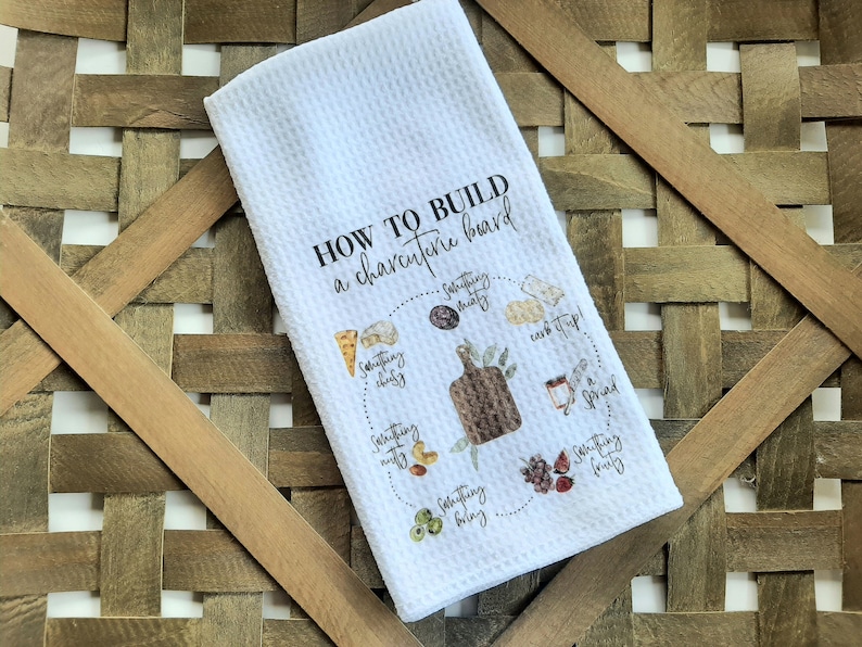 May include: White kitchen towel with a graphic design that says "How to build a charcuterie board" with a circle diagram showing different food items to include on a charcuterie board.