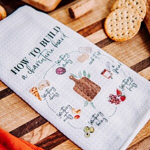 May include: A white kitchen towel with a green and black design that says "How to build a charcuterie board" with a list of items to include on a charcuterie board. The items include something cheesy, something meaty, something nutty, something crunchy, something fruity, and something carb it up!.
