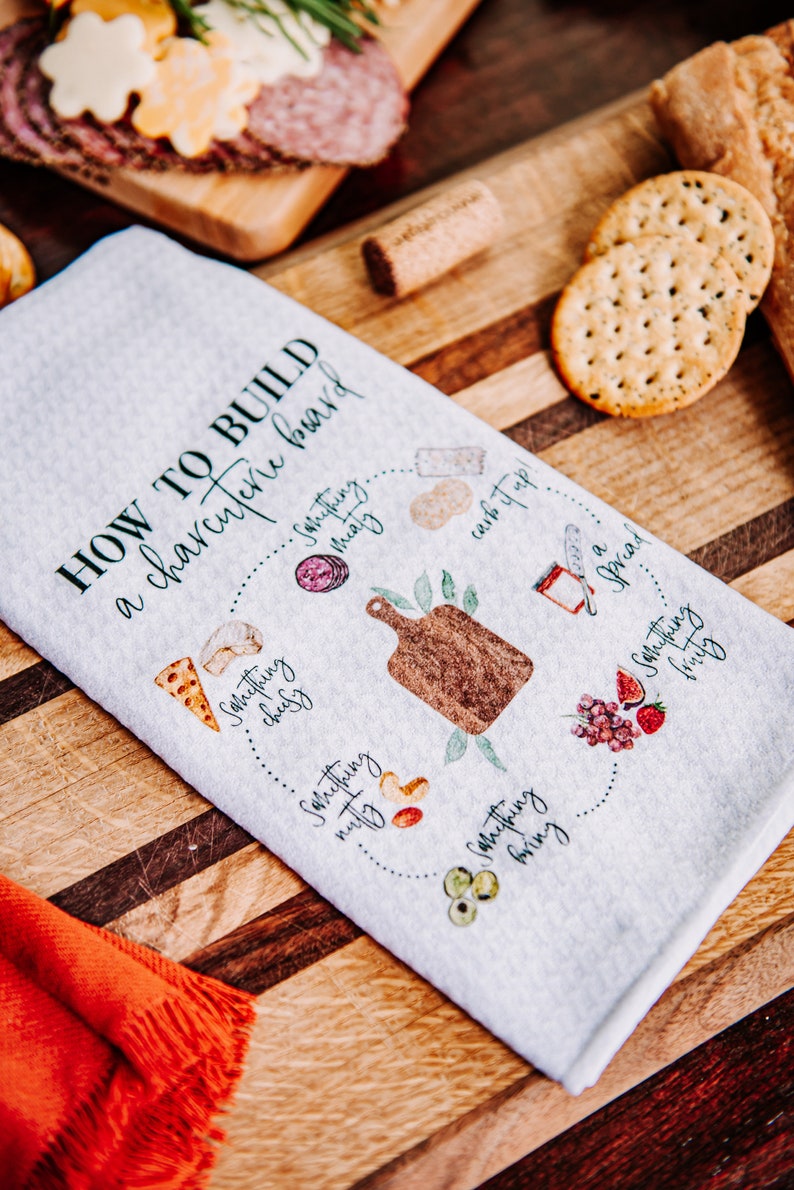 May include: A white kitchen towel with a green and black design that says "How to build a charcuterie board" with a list of items to include on a charcuterie board. The items include something cheesy, something meaty, something nutty, something crunchy, something fruity, and something carb it up!.