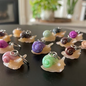 May include: A collection of 11 colorful snail charms with silver keychains. The snails are made of polymer clay and have different colored shells, including purple, pink, green, and brown.