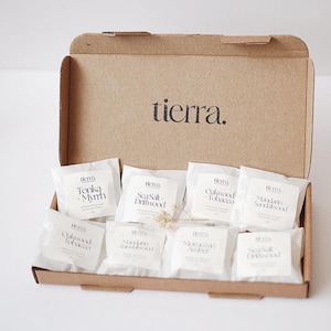 May include: A brown cardboard box with the word "tierra" printed on the lid. Inside are several white packets with scent names such as "Tonka + Myrrh" and "Sea Salt Driftwood".