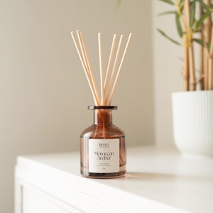 May include: A brown glass diffuser bottle with wooden reeds, labelled "Moroccan Amber." The bottle sits on a white surface, with a plant in a white pot in the background. The diffuser is a home fragrance product.