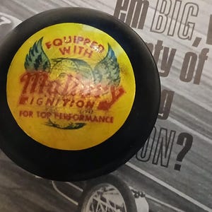 May include: A black, circular automotive part with a yellow center. The center features the text "Equipped with Mallory Ignition for Top Performance" in red and white, with a winged design. The part is set against a black and white background.