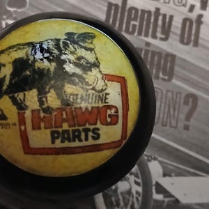May include: A circular black button with a yellow centre. It features a black and white illustration of a pig and the words "GENUINE HAWG PARTS" in red and white.