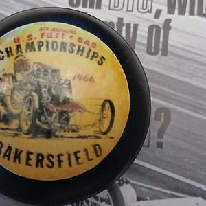 May include: A black, circular object with a yellow centre featuring a vintage drag racing design. The text reads "U.S. Fuel & Gas Championships 1966 Bakersfield". The image is a close-up of a racing-themed collectible.