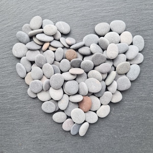 May include: A heart shape composed of smooth, grey river stones. The stones vary in size and shade, with a few tan and reddish-brown stones interspersed. The arrangement is set against a dark grey, textured background.