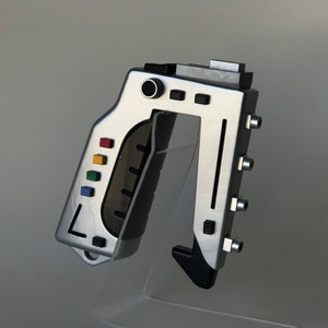 May include: A silver and black handheld device with a black handle and a silver body. The device has a series of coloured buttons and a black trigger. The device is on a clear acrylic stand.