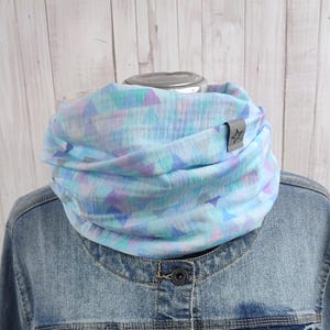 Könnte beinhalten: Ein hellblau-lila Dreiecksmuster-Loop-Schal. Der Schal ist um eine Jeansjacke gewickelt. Ein kleines graues Etikett mit einem Stern ist am Schal befestigt.