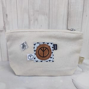 Könnte beinhalten: Eine weiße Canvas-Reißverschlusstasche mit einem braunen Leder-Friedenszeichen-Patch und einem schwarzen Herz-Druck. Die Tasche hat auch zwei kleinere Patches mit Friedenszeichen.