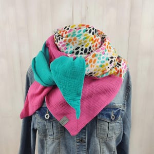 Könnte beinhalten: Ein mehrfarbiger Schal mit auffälligem Design. Der Schal kombiniert Pink, Türkis und einen gemusterten Stoff mit schwarzen, gelben, orangefarbenen und rosa Punkten auf weißem Hintergrund. Der Schal wird über einer Jeansjacke getragen.
