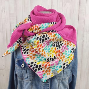Könnte beinhalten: Ein lebendiger, farbenfroher Schal mit einem rosa Rand und einem weißen Hintergrund, der mit bunten Punkten bedeckt ist. Der Schal ist um eine Jeansjacke gewickelt und zeigt sein Design und seine Textur. Das Design des Schals umfasst schwarze, orangefarbene, gelbe und blaue Punkte.