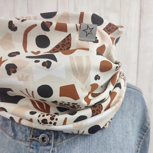 Könnte beinhalten: Ein cremefarbener Rundschal mit einem sich wiederholenden Muster aus schwarzen, braunen und beigefarbenen abstrakten Formen und Leopardenmuster-Akzenten. Ein kleines graues Etikett mit einem Stern ist an der Seite angenäht. Der Schal wird über einer Jeansjacke getragen.