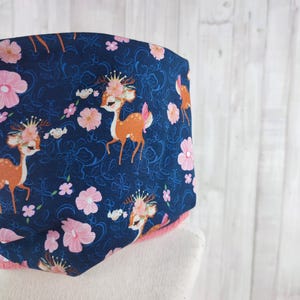 Op de afbeelding: Een donkerblauwe stof met een herhalend patroon van roze bloemen en bruine herten die kronen dragen. De herten kijken in verschillende richtingen en hebben een speelse, cartoonachtige stijl.