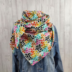 Könnte beinhalten: Ein weißer Schal mit einem bunten abstrakten Muster aus schwarzen, orangen, gelben, rosa und blauen Punkten. Der Schal ist gefaltet und um den Hals einer Person gelegt.