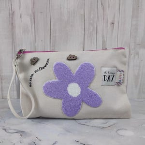 Op de afbeelding: Een witte canvas clutch met een paarse pluche bloemenapplicatie, twee houten wolkvormige knopen en de tekst "no rain. no flowers." en "oh happy DAY" aan de voorkant.