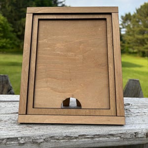 Könnte beinhalten: Ein hellbrauner Holzbilderrahmen. Der Rahmen hat eine rechteckige Öffnung und einen kleineren rechteckigen Rand im Inneren. Unten befindet sich ein halbkreisförmiger Ausschnitt. Der Rahmen steht auf einer Holzoberfläche.