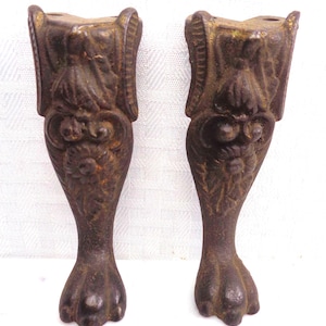 May include: Two dark brown metal furniture legs featuring a claw and ball foot design. Each leg has an ornate, raised floral and leaf pattern along the upper section and two circular mounting holes at the top for attachment to a base.