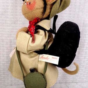 May include: A tan felt mouse doll wearing a beige trench coat, a green hat, and a red bandana. The mouse carries a black backpack and a green canteen. A white tag on the backpack reads Annalee 2004. The doll has tan shoes with brown accents.