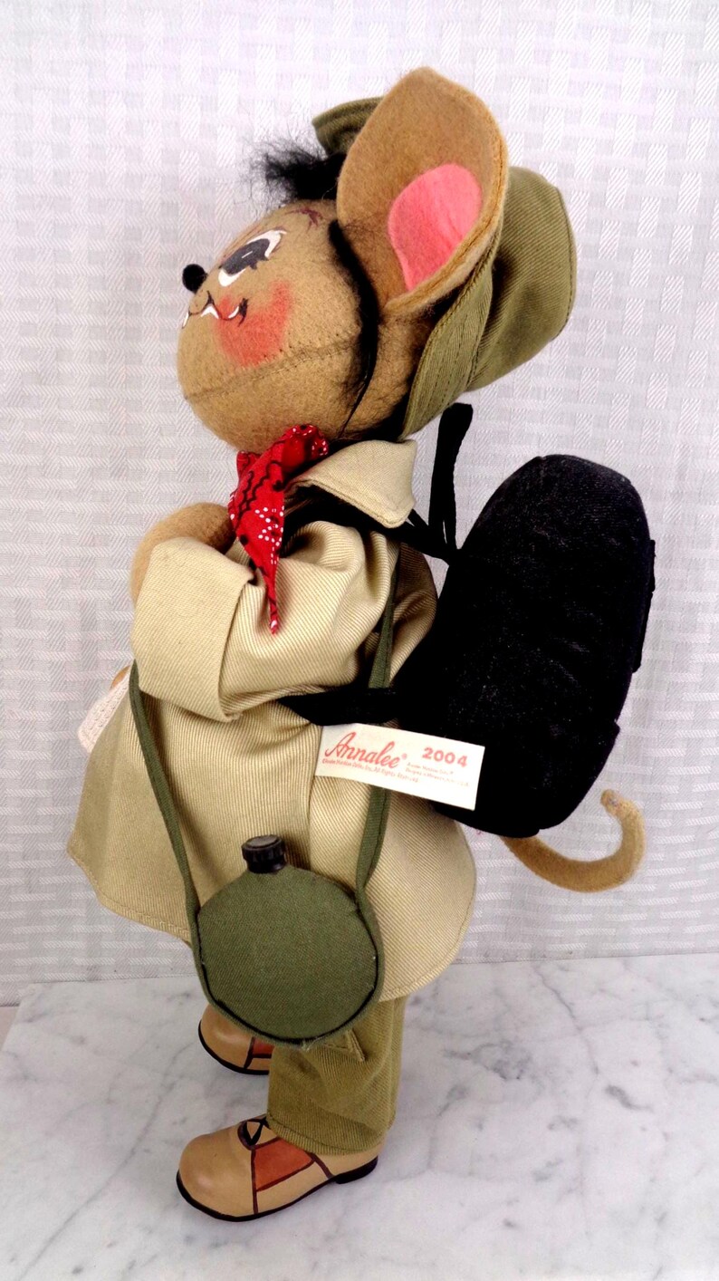 May include: A tan felt mouse doll wearing a beige trench coat, a green hat, and a red bandana. The mouse carries a black backpack and a green canteen. A white tag on the backpack reads Annalee 2004. The doll has tan shoes with brown accents.