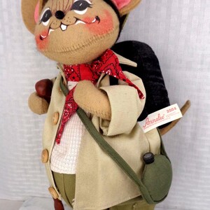 May include: A felt mouse doll wearing a tan jacket, olive green pants, a red bandana, and a green hat. The mouse carries a black backpack, a green canteen, and a brown walking stick. A white tag on the backpack reads Annalee 2004.