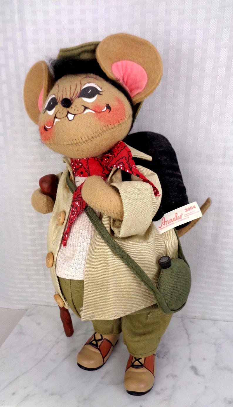 May include: A felt mouse doll wearing a tan jacket, olive green pants, a red bandana, and a green hat. The mouse carries a black backpack, a green canteen, and a brown walking stick. A white tag on the backpack reads Annalee 2004.