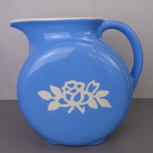 May include: A light blue ceramic pitcher with a white rose design. The pitcher has a round body and a handle.