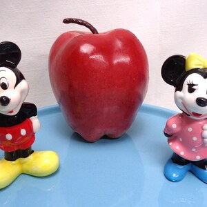 May include: A red apple sits between two ceramic figurines of Mickey and Minnie Mouse. Mickey wears a red and yellow outfit, while Minnie is in a pink polka-dot dress with a yellow bow. The figures are on a light blue surface.
