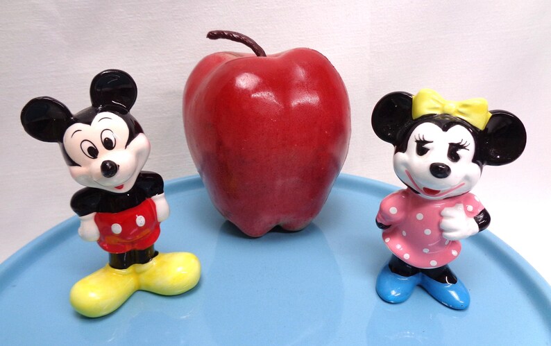 May include: A red apple sits between two ceramic figurines of Mickey and Minnie Mouse. Mickey wears a red and yellow outfit, while Minnie is in a pink polka-dot dress with a yellow bow. The figures are on a light blue surface.