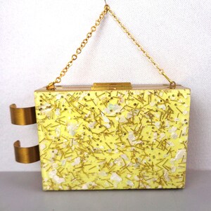 May include: A rectangular yellow clutch with gold accents and a chain strap. The clutch is covered in gold and white glitter and has two gold metal rings on the side. The clasp and chain are also gold.