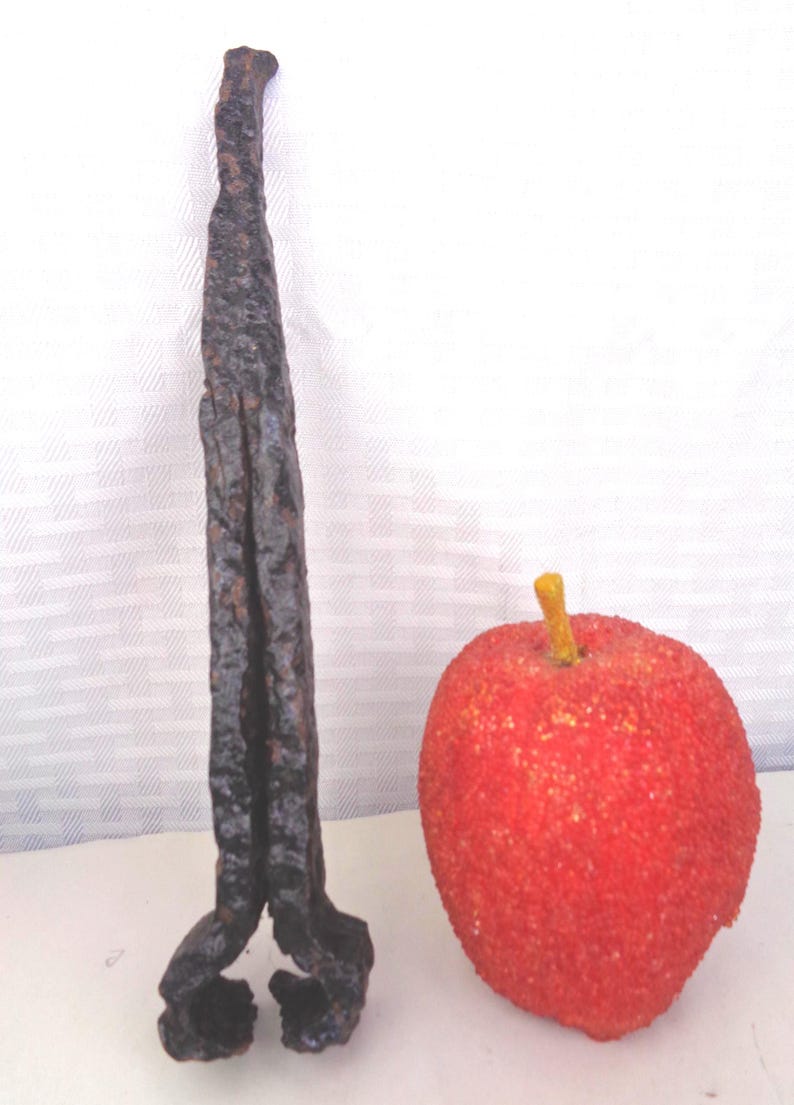 May include: A dark, textured metal object with a twisted design stands next to a red, textured apple with a yellow stem. The metal object has a rough, aged appearance, while the apple is round and vibrant.