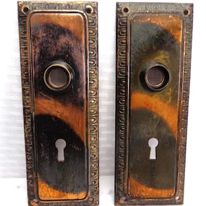 May include: Two antique door handle backplates with a rectangular shape and ornate trim. The plates have a dark, aged appearance with bronze and black tones. Each plate features a keyhole and a circular knob.