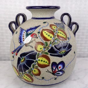 May include: A beige ceramic vase with a round body and two handles. The vase is decorated with a colorful bird, flowers, and a butterfly. The rim and handles are dark blue.