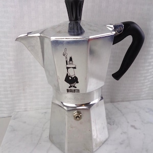 May include: A silver aluminum stovetop espresso maker with a black plastic handle and lid knob. The body features a black graphic of a man with a mustache pointing one finger upward, with the text BIALETTI and Moka Express printed below.
