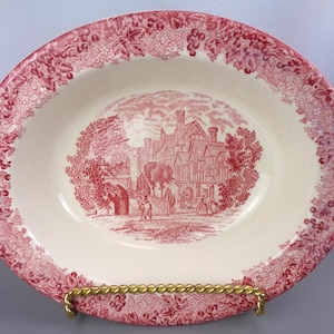 May include: Oval ceramic serving dish with a red and white design. The rim features a grape vine pattern, and the center depicts a detailed red illustration of a building and figures. The dish is displayed on a gold-colored metal stand.