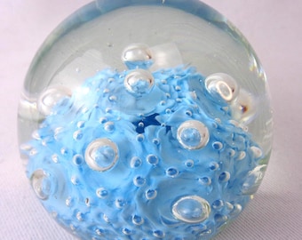 May include: A spherical glass paperweight featuring a clear outer layer encasing a textured, light blue interior core. The design includes numerous trapped air bubbles of varying sizes distributed throughout the clear glass sphere.